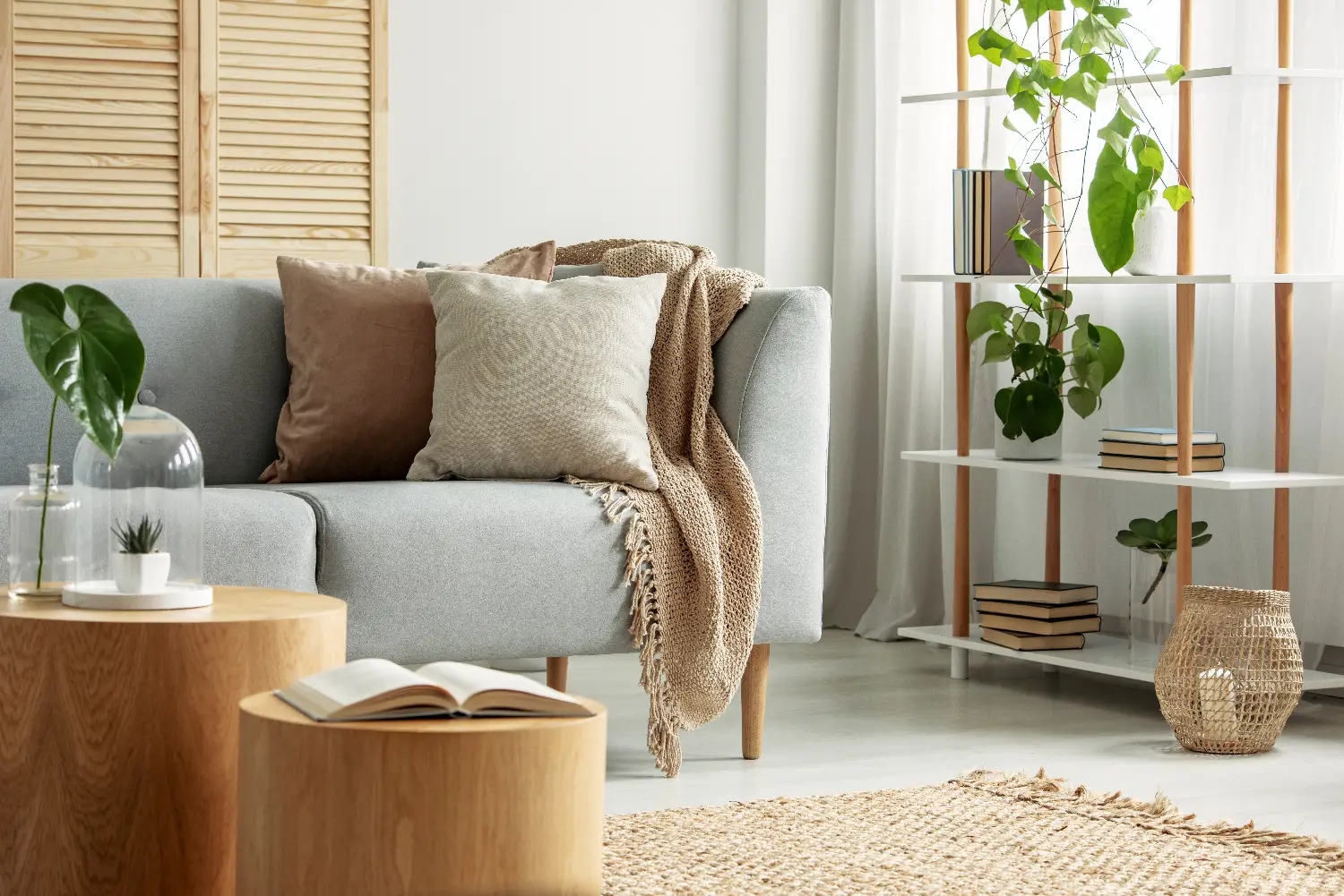 Der Einblick ins Wohnzimmer Wohnzimmer zeigt ein Sofa mit zwei Polstern und einer Decke sowie ein Regal mit einigen Büchern und einer Pflanze. Im Vordergrund stehen zwei kleine runde Couchtische aus Holz mit einer Pflanze und einem Buch darauf.